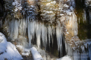 Im Sonnenlicht wirken die Eiszapfen vor den kleinen Höhlungen des Wasserfalls noch schöner